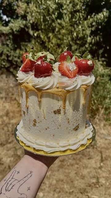 Hand holding a white strawberry celebration cake with gold details outdoors